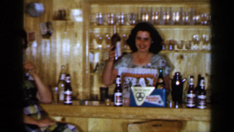 1965 Long Island frame showing a woman smiling behind a wood-paneled bar with bottles, glassware, and a branded-looking box nearby.