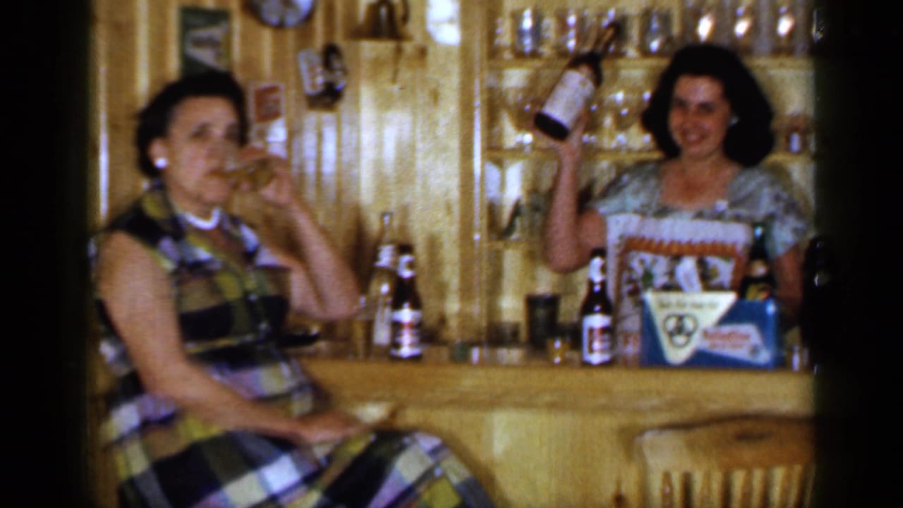 Two women at a wood-paneled bar with drinks and bottles on the counter.