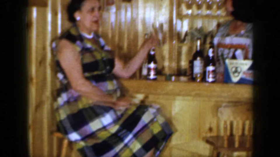 1965 Long Island frame of a woman seated at a wood bar, with bottles, glass shelves, and a partial figure behind the counter.