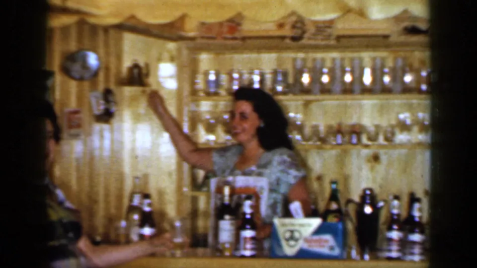 1965 Long Island home-movie frame with a woman behind a wood-paneled bar lifting a bottle near rows of glassware and drinks.
