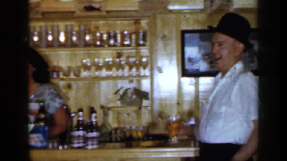 1965 Long Island frame showing a man in a dark hat holding a drink near a wood-paneled bar with bottles and glassware.