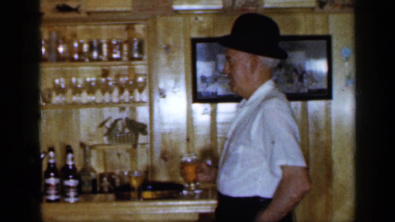 Older man in a black bowler hat holds a beer glass beside a wooden bar.