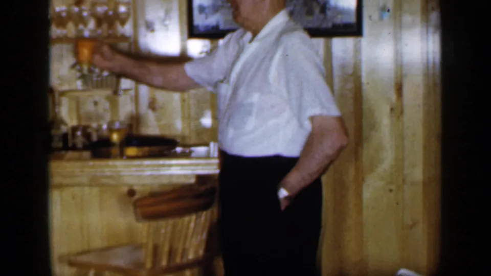 The man lifts a beer glass in a wood-paneled bar interior, with shelves of drinking glasses, bottles, and a framed picture visible behind him.