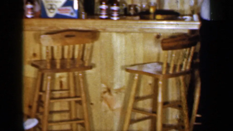 A low view of the wooden bar shows stools, bottles, shelves, and part of a standing figure at the edge of the frame.