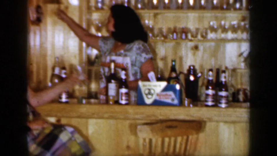 A woman behind the wooden bar reaches upward near shelves of glasses, with bottles and a visible beer carton or branded packaging on the counter.