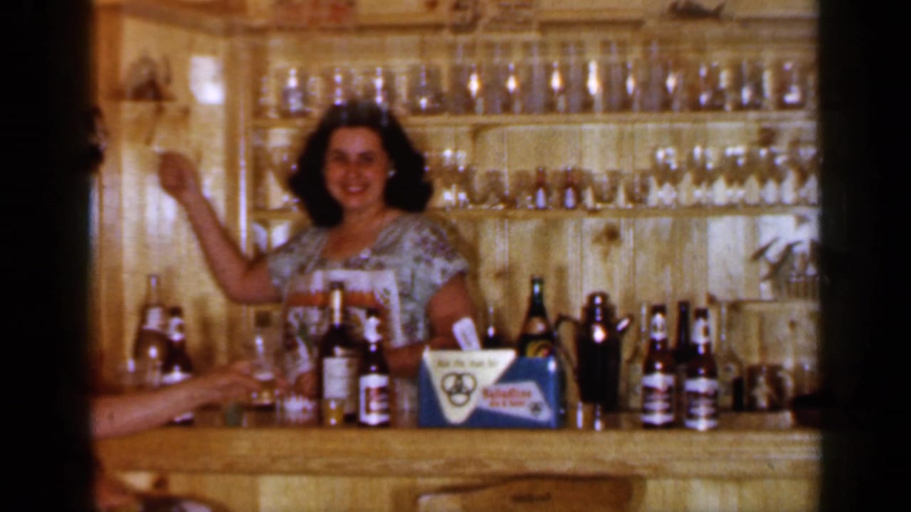1965 Long Island home-movie frame showing a woman behind a wooden bar with bottles, glasses, and a partially visible adult nearby.