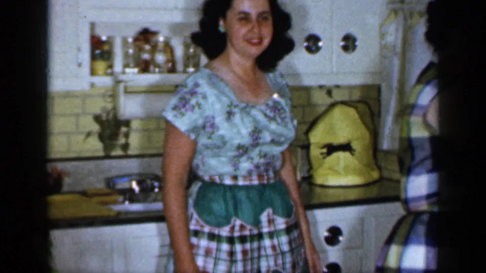 1965 Long Island kitchen scene with a woman in a floral blouse and green apron standing near white cabinets, a sink, and tiled backsplash.