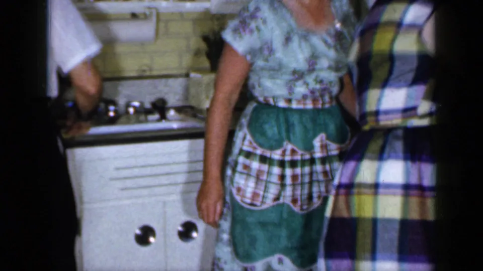 Cropped 1965 Long Island home-kitchen frame showing a woman in an apron beside another adult in a plaid dress near the sink and cabinets.