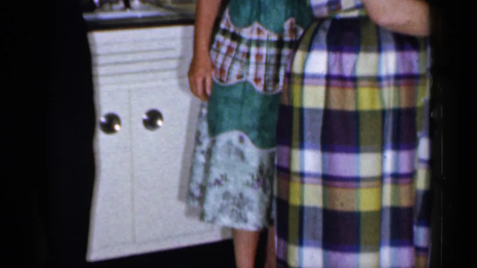 1965 Long Island home-movie frame with partial views of two adults in a kitchen, including plaid clothing, white cabinets, and a lace-trimmed apron.