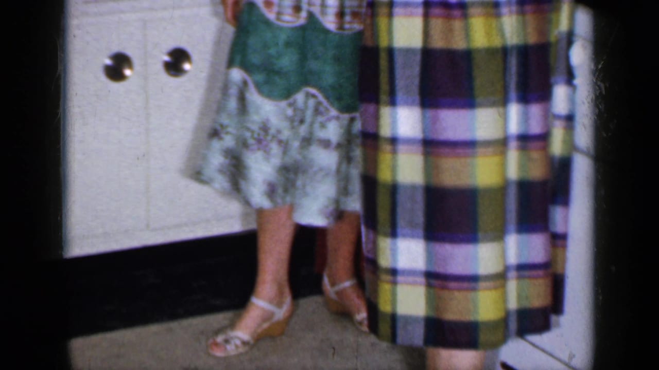 1965 Long Island home-movie frame showing women's dresses and sandals beside a white cabinet in a private kitchen.