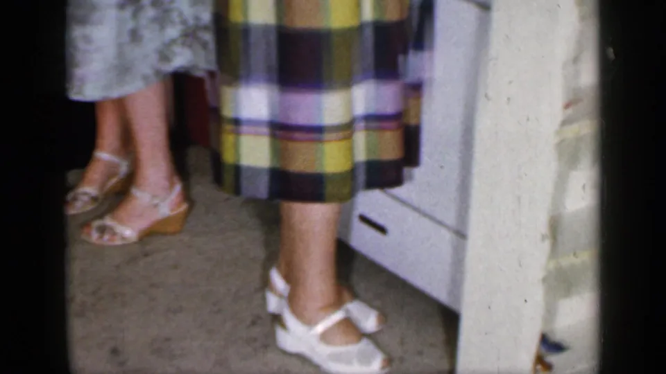 1965 Long Island domestic footage frame focused on women's shoes and skirt hems beside a kitchen counter and white appliance.