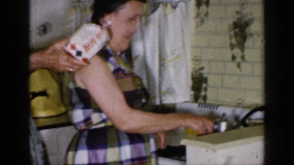 1965 Long Island kitchen frame showing a woman in plaid holding a boxed item and reaching toward the counter near a tiled wall.