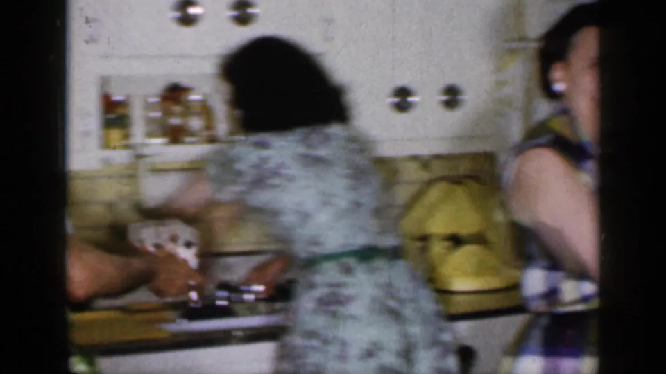 1965 Long Island kitchen scene with a woman in a floral dress moving at the counter and another woman partly visible near white cabinets.