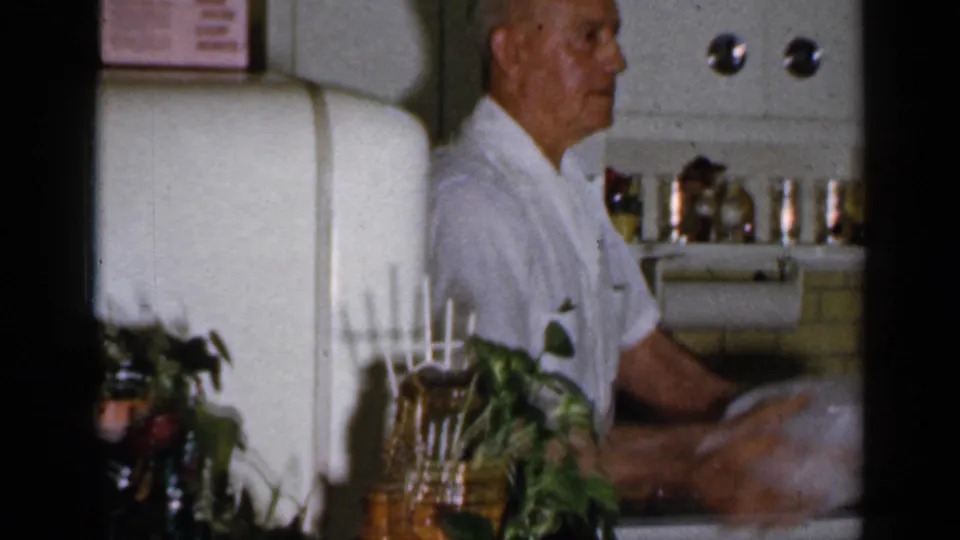 Older man in a white shirt appears at the counter in a 1965 Long Island kitchen with refrigerator and shelves behind him.