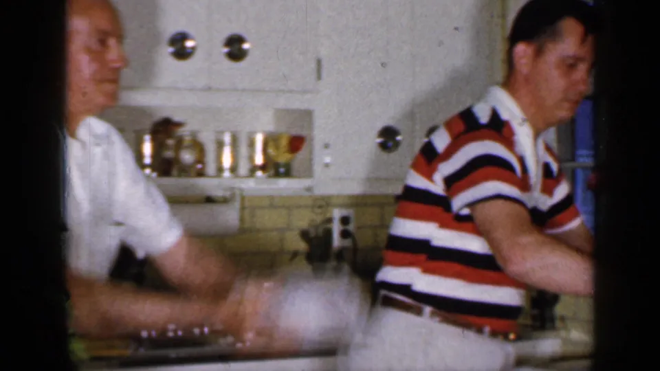 Two men gather at the kitchen counter in the Long Island home-movie scene, with cabinets and shelf items visible behind them.