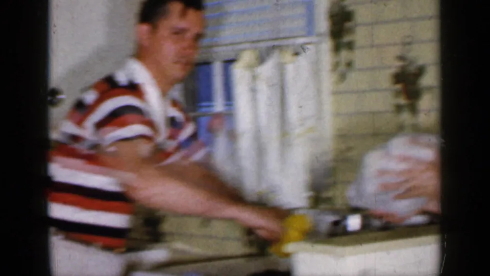 A younger man in a striped polo reaches at the counter in the 1965 Long Island kitchen, with window, towels, and sink area visible.