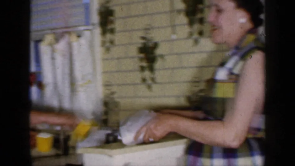 The woman continues handling dough in the Long Island kitchen, with yellow tile, a curtained window, and hanging plants visible.
