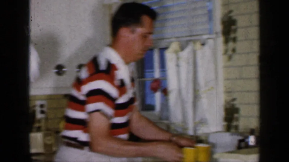 A younger man in a striped shirt joins the kitchen activity near the counter, yellow cup, curtained window, and tile wall.