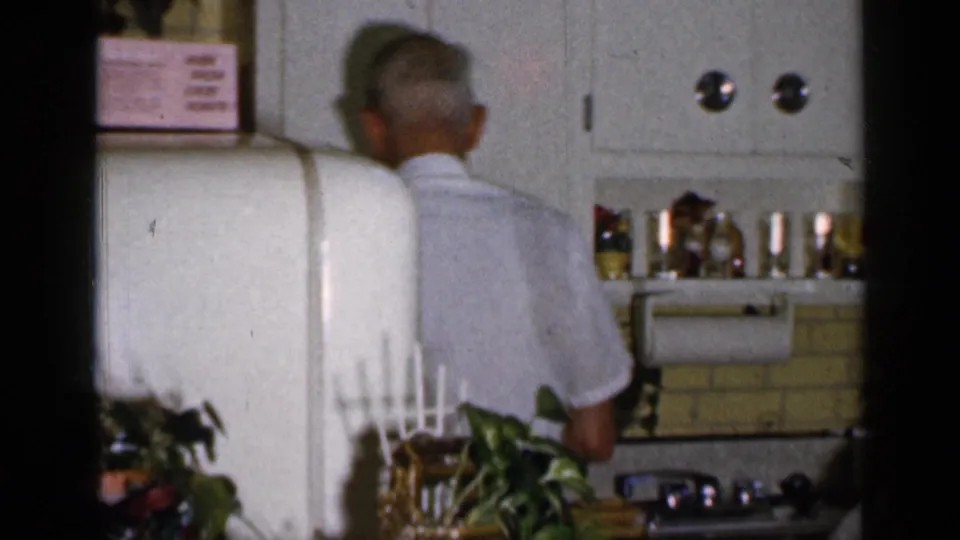 An older man stands at the counter beside a white refrigerator, cabinets, utensils, and bottles in the private kitchen.