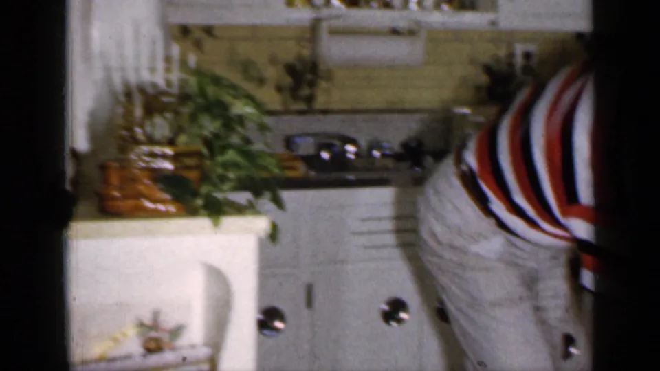 In a 1965 Long Island home kitchen, a man in a red, white, and black striped shirt bends near a white oven and counter plants.