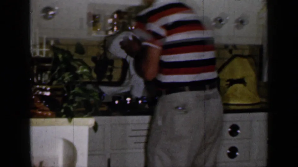 The man stands beside the stove and counter, holding a white cloth in the 1965 Long Island kitchen interior.