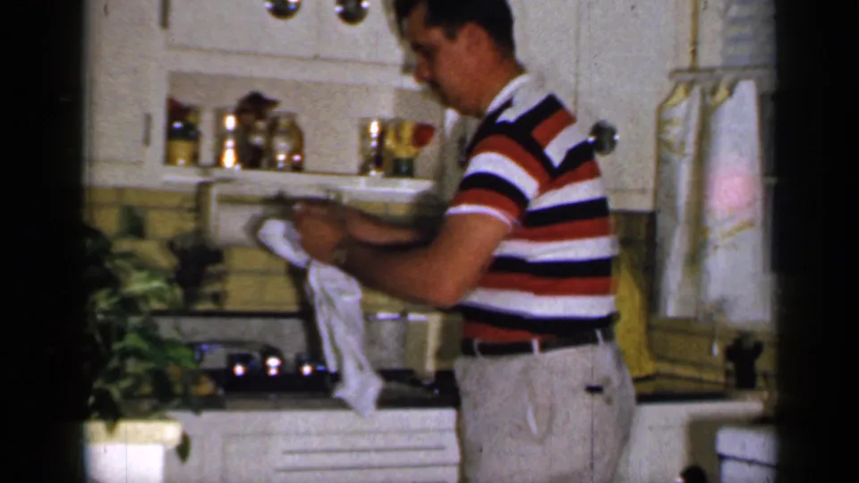 A man in a striped polo dries a dish with a white cloth near the sink, white cabinetry, and yellow backsplash in a 1965 home kitchen.