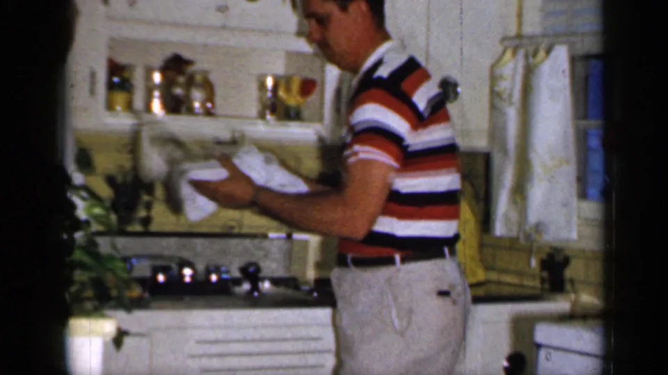 The man continues drying a dish at the counter in the Long Island kitchen, with white cabinets and yellow tile behind him.
