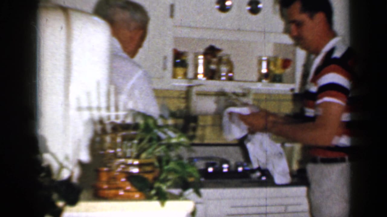Man drying dishes beside an older man in a vintage kitchen.