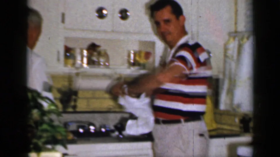 A man in a striped polo turns toward the camera while holding a towel in a 1965 Long Island kitchen interior.
