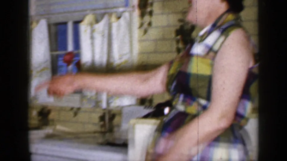 A woman in a colorful plaid dress pours from a kettle near cafe curtains and yellow tile in the Long Island kitchen.