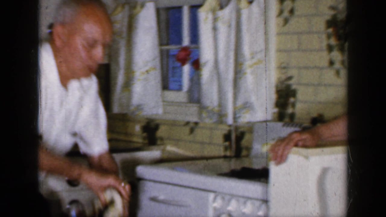 Adult man in a white shirt wipes or handles something near a kitchen sink.