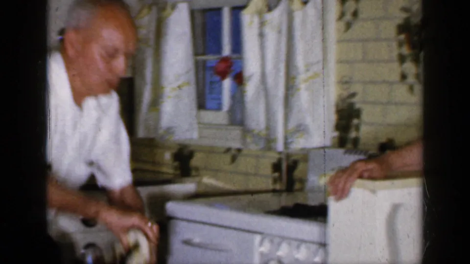 In a 1965 Long Island home kitchen, an adult man in a white short-sleeved shirt works near the sink and tiled wall.