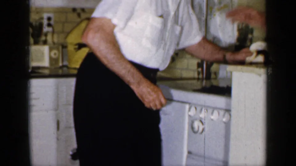 An adult man in a white shirt wipes or reaches across a counter in a 1965 Long Island kitchen interior.