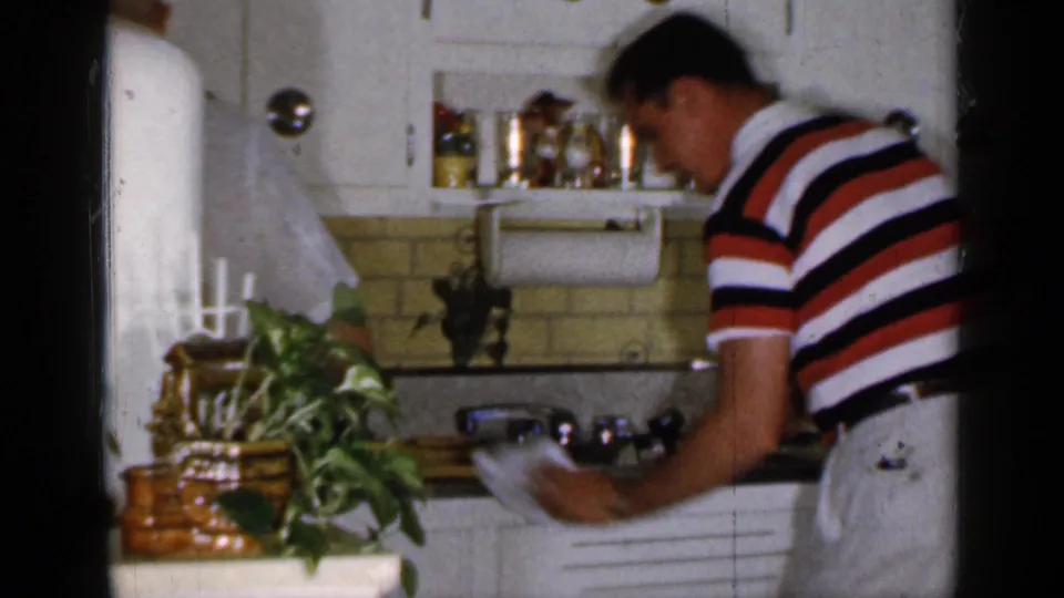 A man in a red, white, and dark striped polo washes dishes at the sink in a 1965 Long Island kitchen.