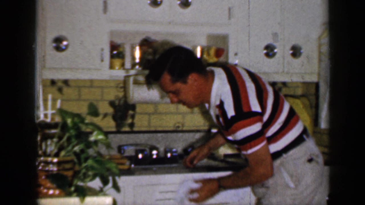 Man in a striped shirt bends over a kitchen counter with white cabinets and yellow tile behind him.