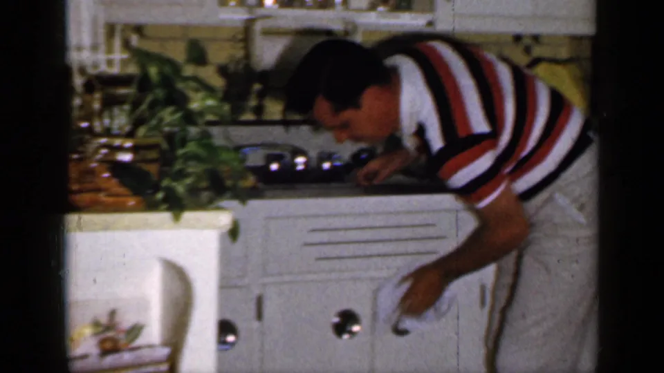 A man in a striped polo appears to wipe the kitchen counter in a 1965 Long Island interior, with white cabinets and a stove visible.