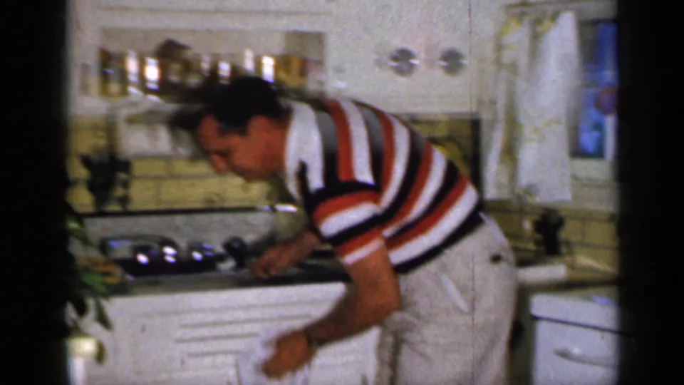 The man continues cleaning the kitchen counter in the 1965 Long Island home-movie footage, partly blurred by motion and film softness.