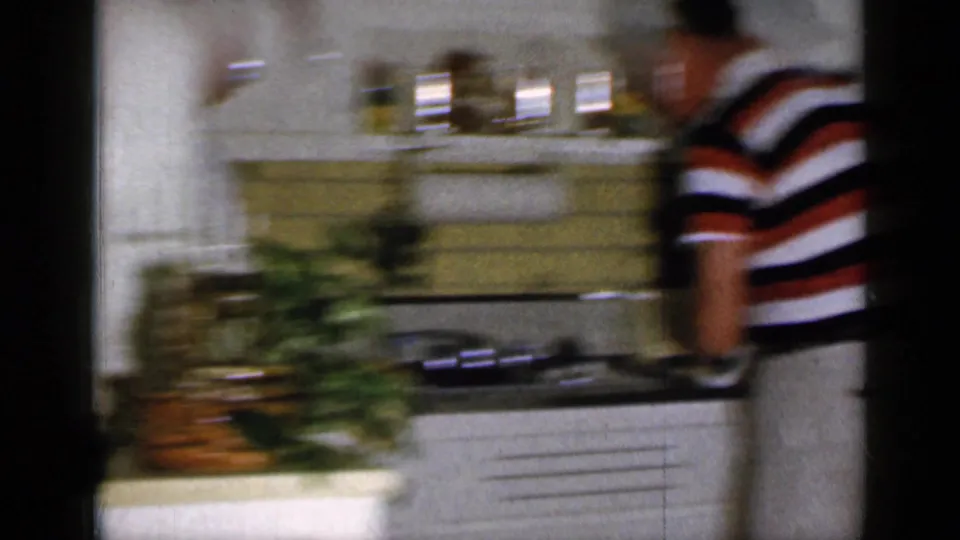 A motion-blurred 8mm frame shows the striped-shirted man moving beside the kitchen counter in the same 1965 Long Island interior.