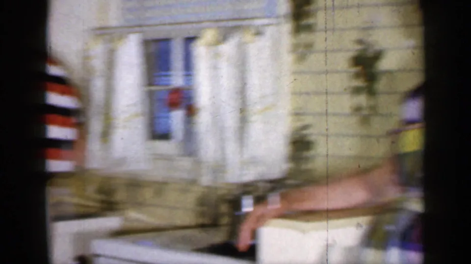 1965 Long Island home-movie frame showing a woman in a colorful plaid dress moving through a kitchen near a sink and curtained window.