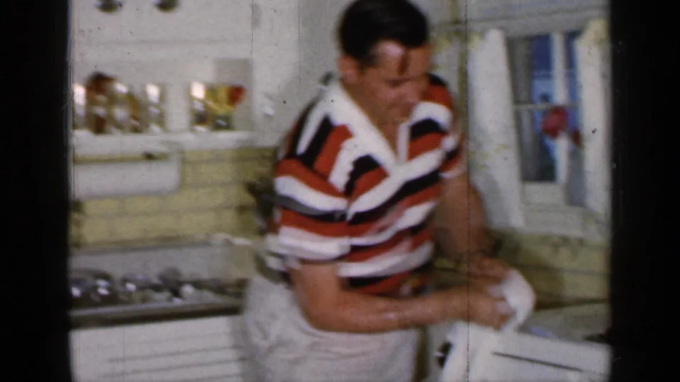 1965 Long Island home-movie frame showing a man in a red, white, and black striped polo washing or drying dishes in a kitchen.