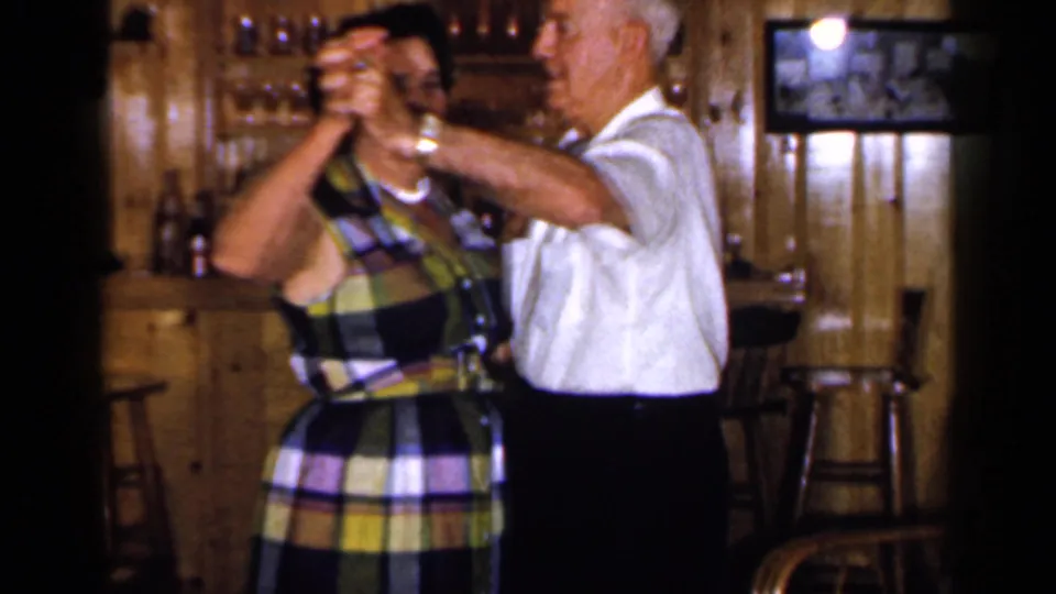 1965 Long Island home-movie frame showing an adult couple dancing in a wood-paneled room with bar area and stools behind them.