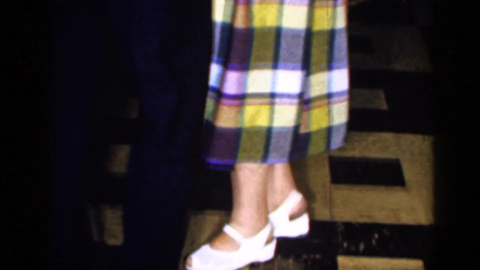 The woman’s plaid dress and white shoes are visible as she dances with a man in the 1965 Long Island bar scene.