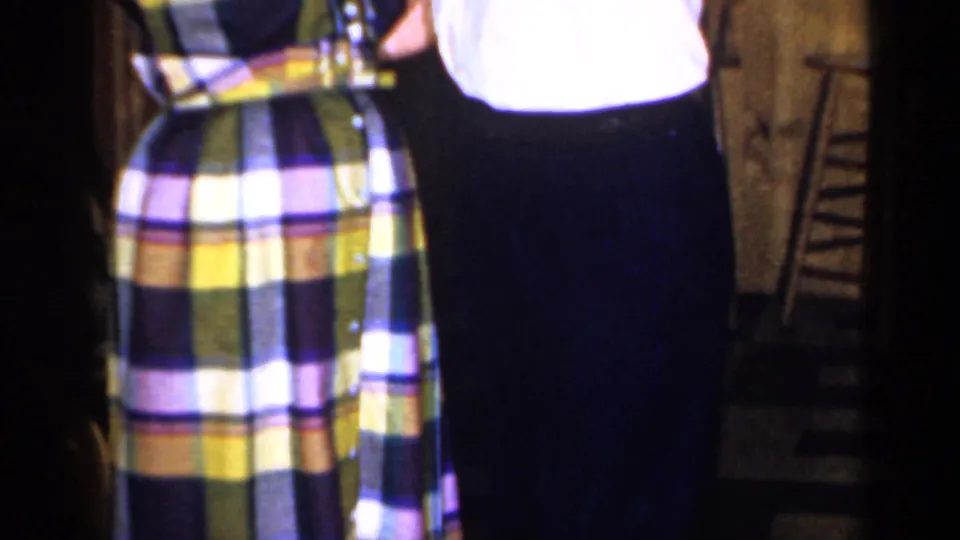Close 1965 home-movie frame of the dancing couple's clothing, with a plaid dress, white shirt, and dark trousers in a private interior.