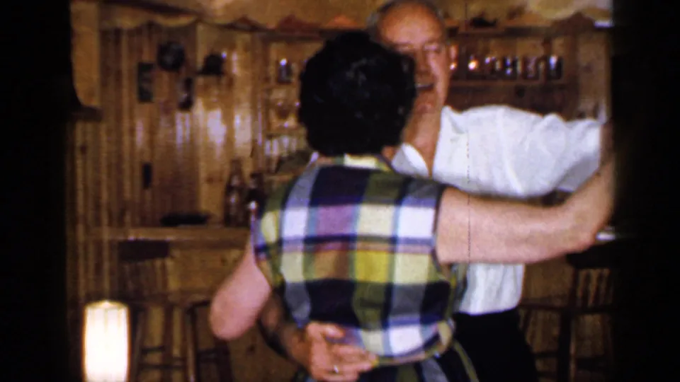 1965 Long Island home-movie frame of an adult couple dancing closely before a wood-paneled bar with shelves, stools, and warm lighting.