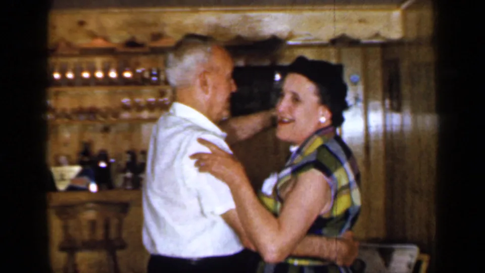 1965 Long Island frame showing a smiling adult couple ballroom dancing beside a wood-paneled bar, with glassware and stools in the background.
