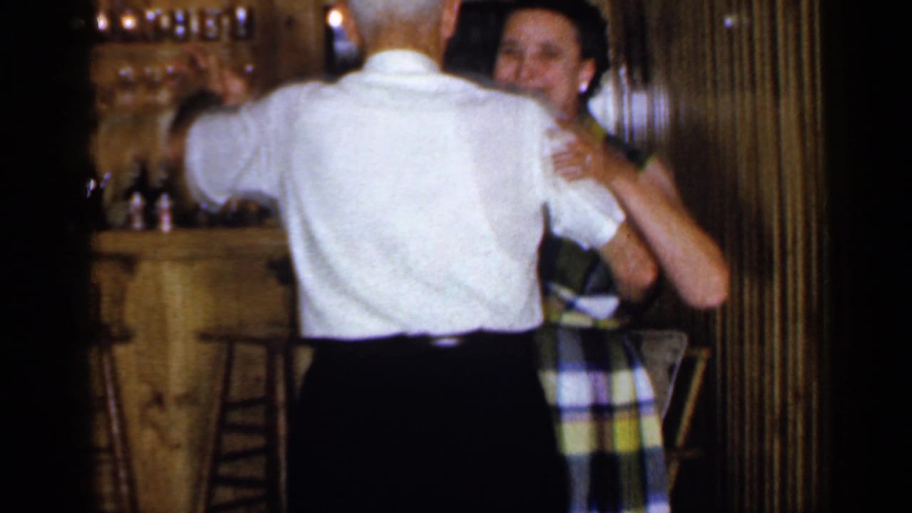 In a 1965 Long Island home-movie frame, an older couple dances near a bar area in a warm wood-paneled interior.
