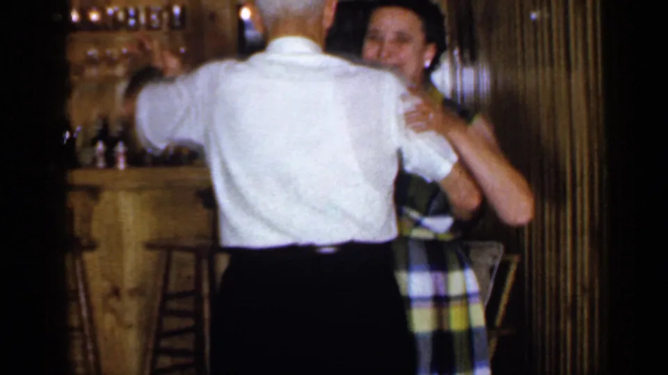 In a 1965 Long Island home-movie frame, an older couple dances near a bar area in a warm wood-paneled interior.