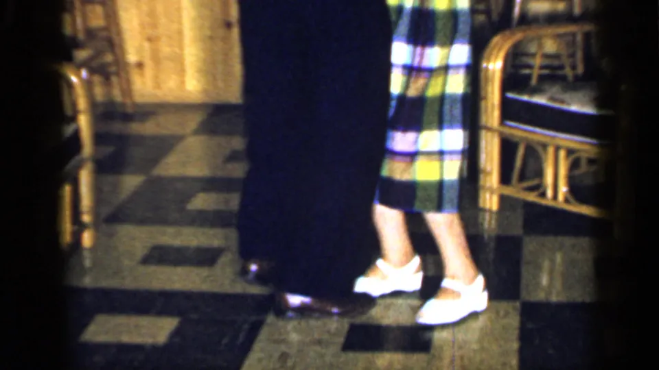 An older couple dances full length on a checkered floor, with the woman in a plaid dress and the man in a white shirt.