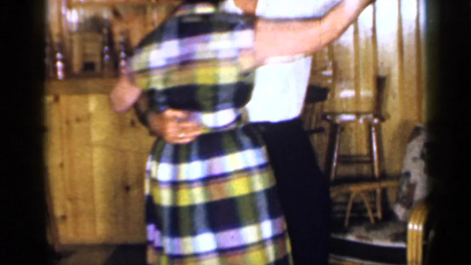 A close, soft 8mm frame shows the older couple mid-dance, with the plaid dress prominent against the wood-paneled room.