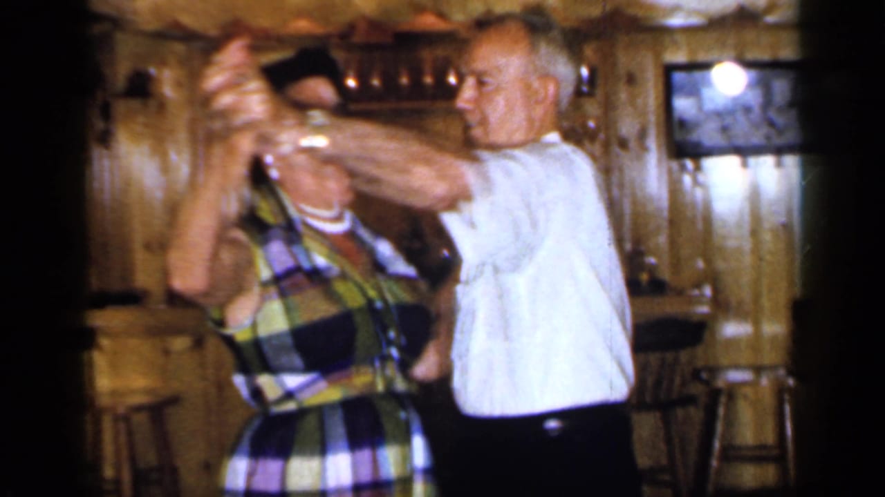 Older man in a white shirt dances closely with a woman in a plaid dress inside a wood-paneled bar.